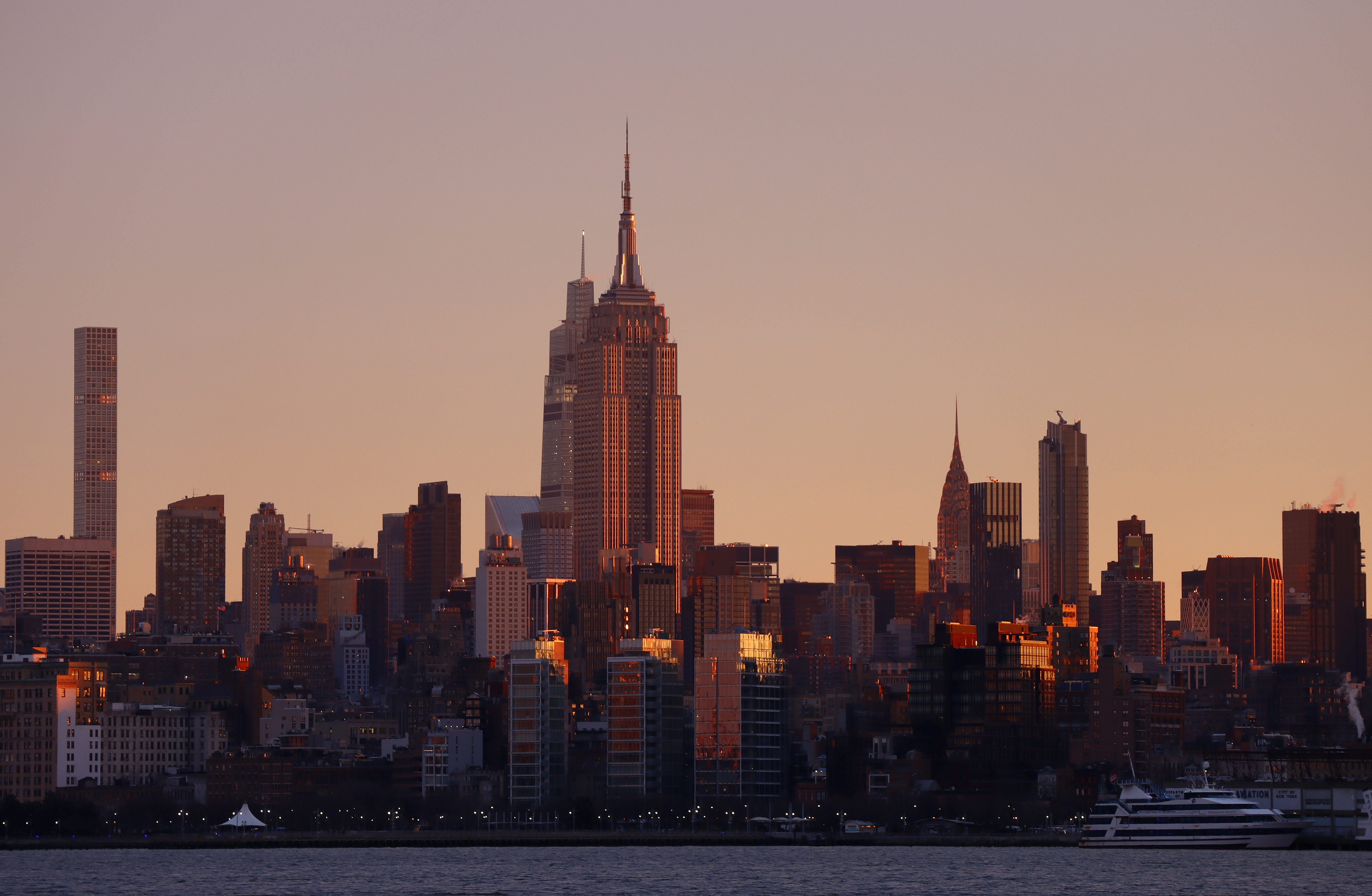 The sun rises behind the skyline of midtown Manhattan, the Empire State Building, One Vanderbilt and the Chrysler Building in New York City