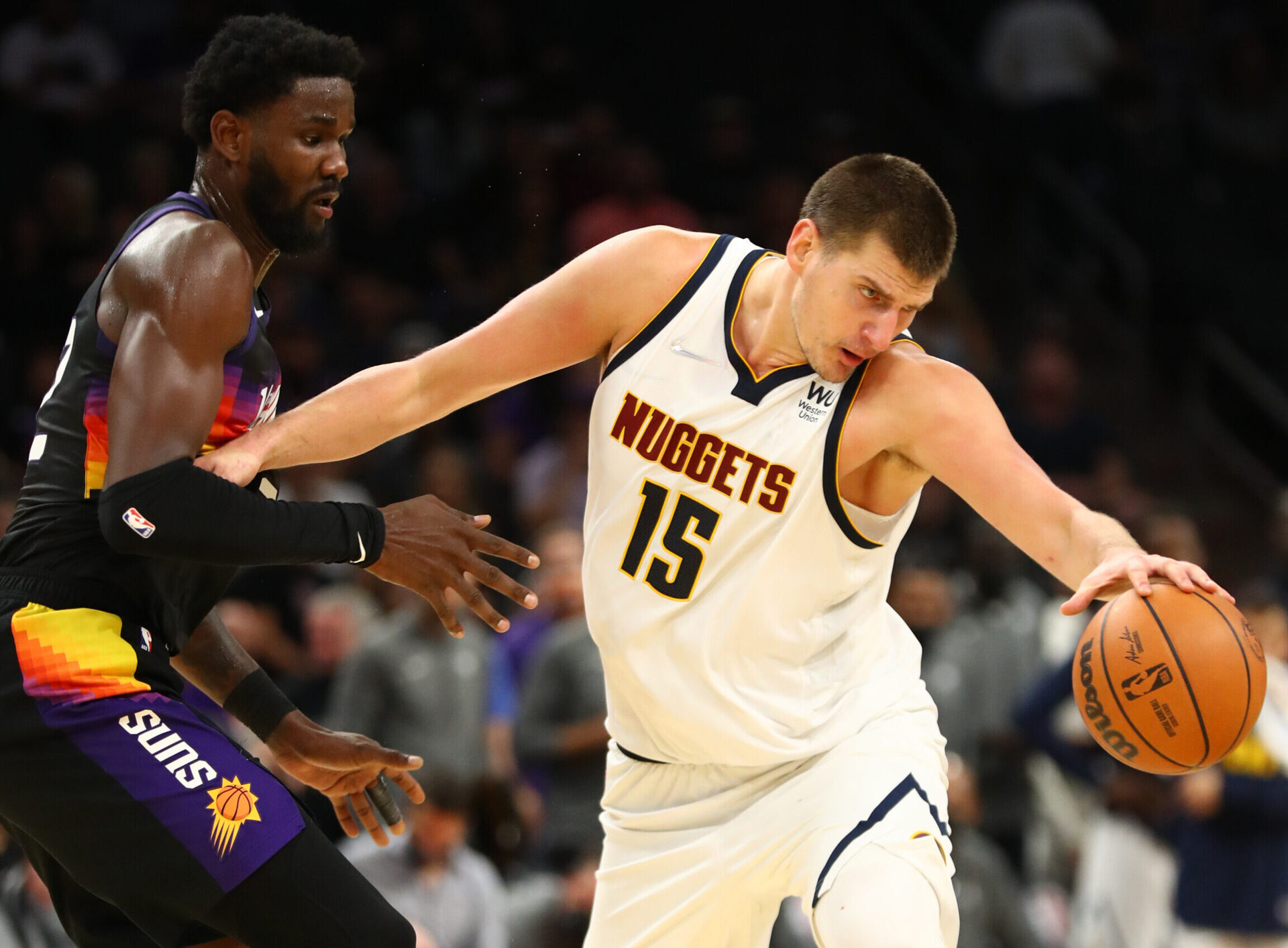 Denver Nuggets center Nikola Jokic (right) bounces the basketball with his left hand while using his right arm to hold off defending Phoenix Suns center Deandre Ayton (left)