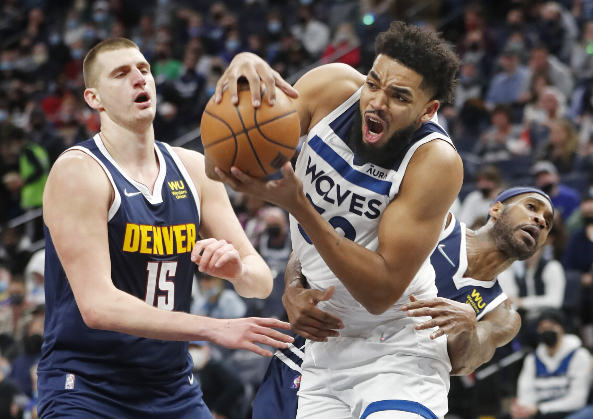 Minnesota Timberwolves center Karl-Anthony Towns (right) grimaces as he tries to control the basketball with both hands while being defended by Denver Nuggets center Nikola Jokic (left) and guard Will Barton (right)