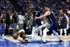 Utah Jazz guard Donovan Mitchell, left, tries to pass the ball while lying on his back, with the Dallas Mavericks' Dorian Finney-Smith, center, and Dwight Powell, right, standing over him on Monday, April 25, 2022, in Dallas.